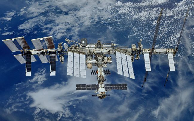 The International Space Station flying above a blue, cloudy Earth below. It is an assortment of long, rectangular solar panels, silver cylinders and other metallic structures that point out at odd angles. (Credit: NASA/Roscosmos)